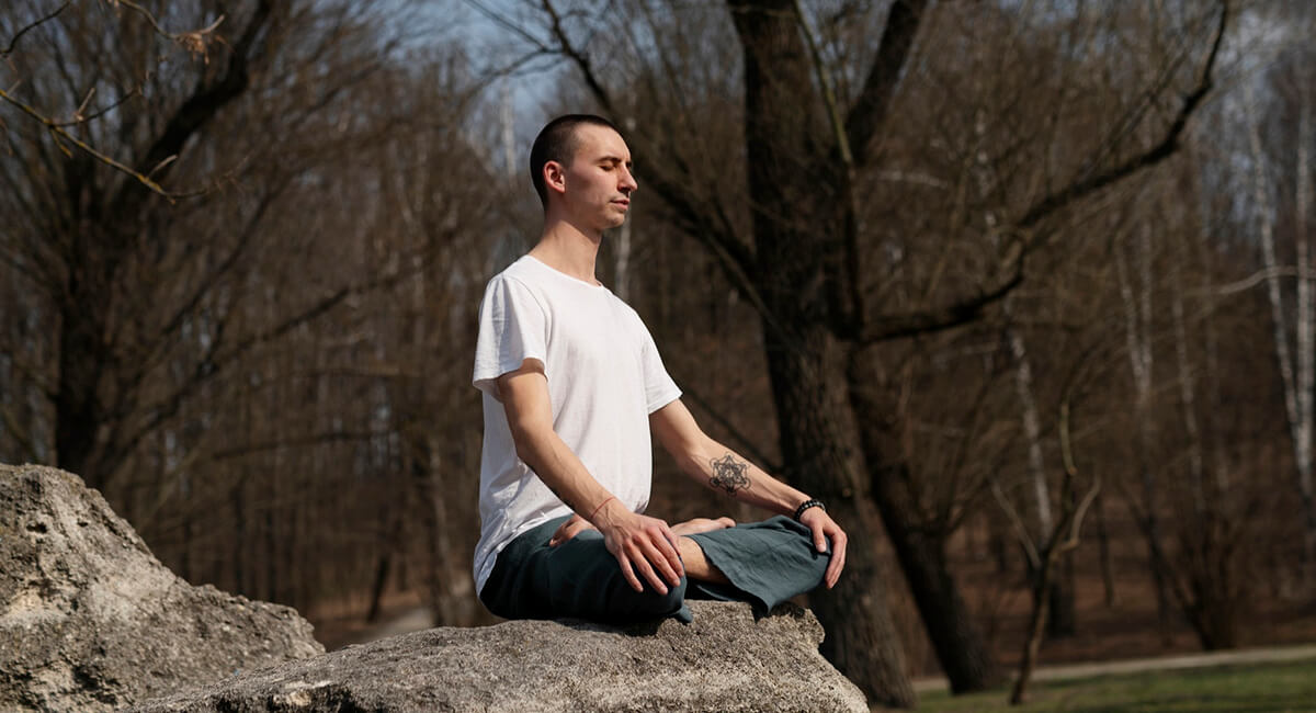 A man doing yoga while sitting on top of a natural stone.