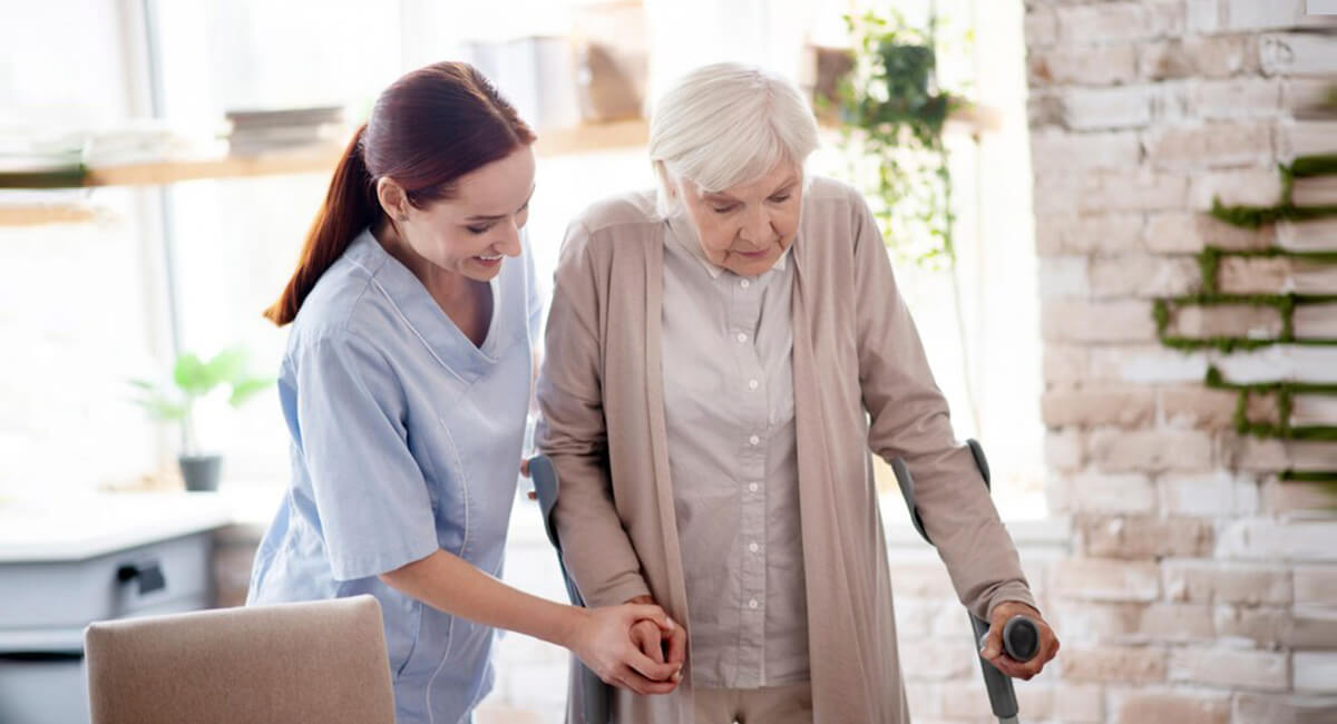 A carer providing assistance to an elderly woman using crutches