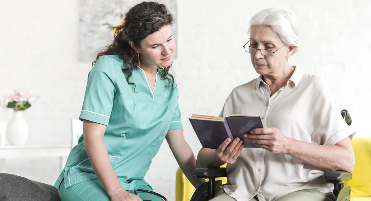 A carer assisting an elderly woman in a wheelchair to read a book.