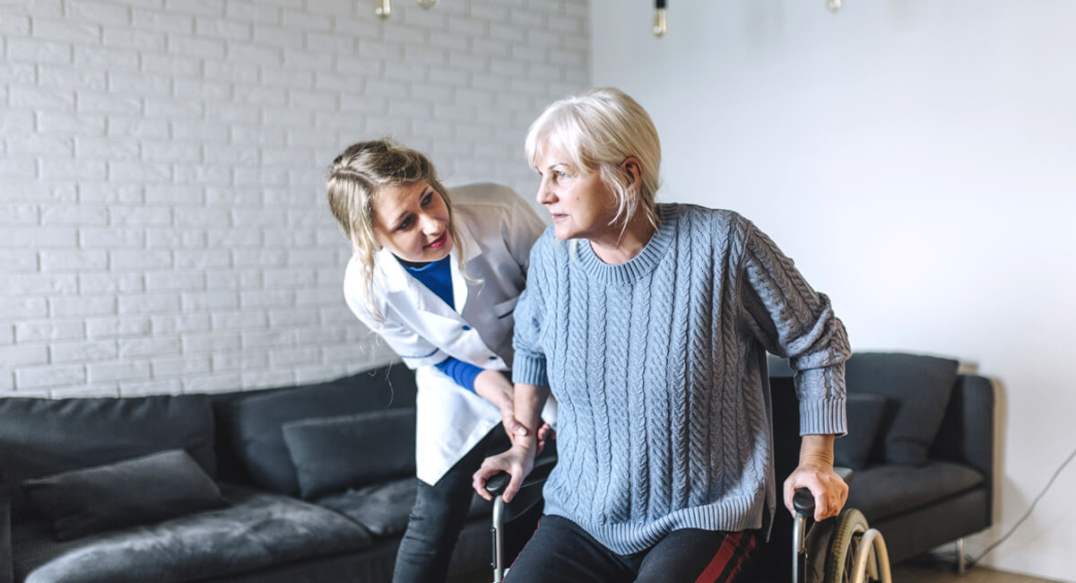 Assisted by a carer, an elderly woman tries to get up from her wheelchair.