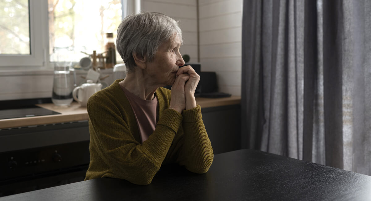 An elderly woman seated in her kitchen and staring out of her window