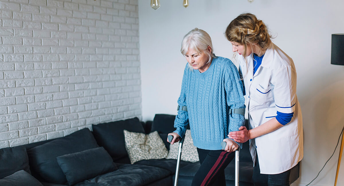 An in-home carer walks an elderly woman with her walking crutches.